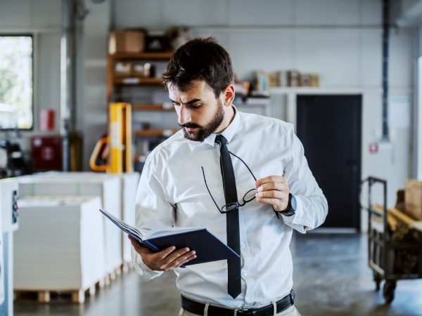 Handsome caucasian serious bearded graphic engineer in shirt and tie, holding notebook and looking at it while standing in printing shop.